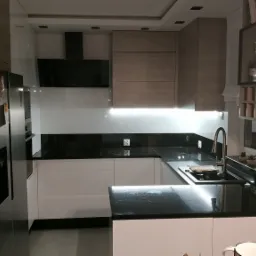 Nowoczesna kuchnia z białymi frontami szafek i czarnym blatem, oświetlenie LED pod szafkami wiszącymi, widoczny fragment lodówki i szafka z ekspozycją na butelki.