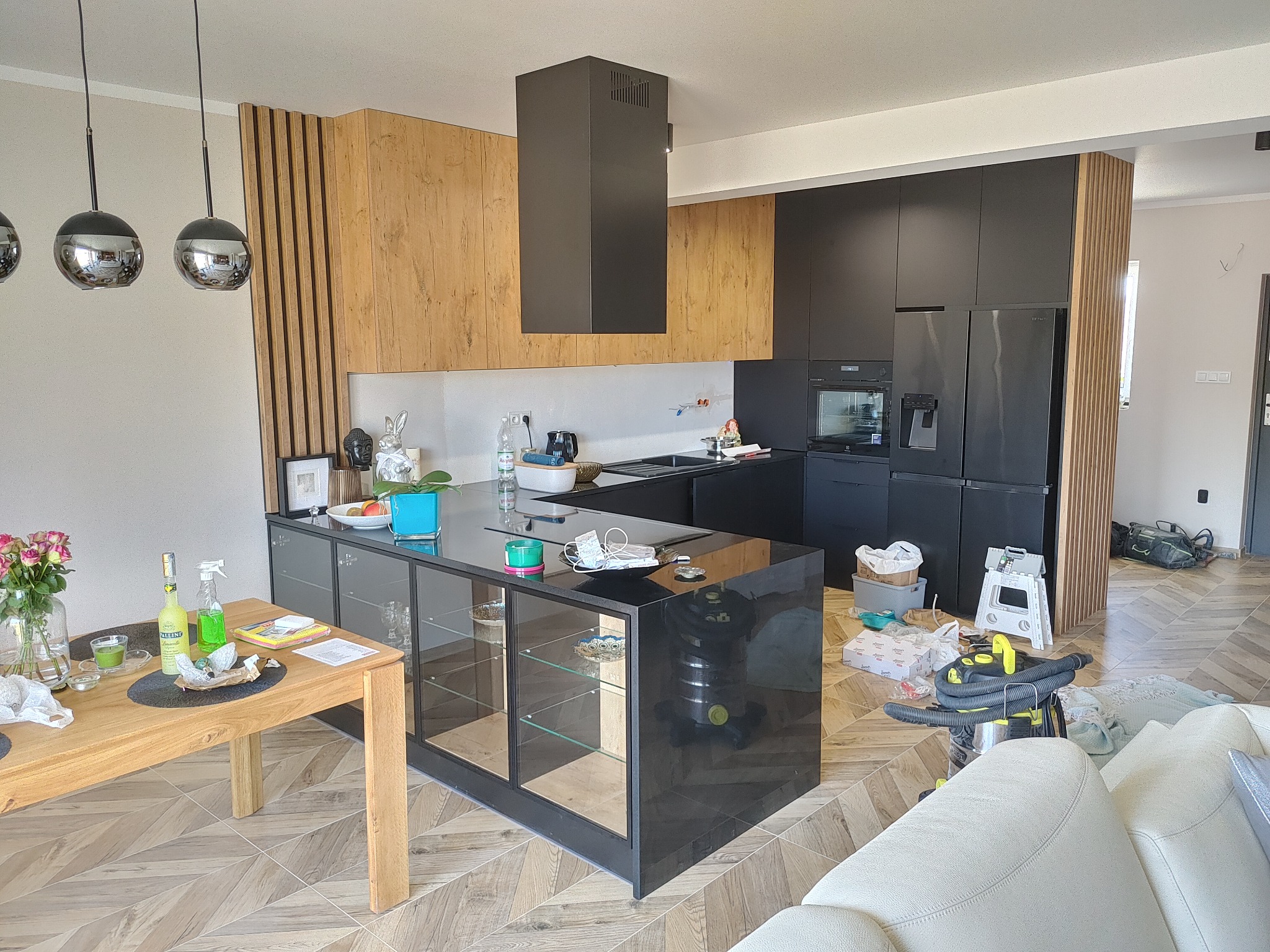 Nowoczesna kuchnia z zabudową w kolorach drewna i czerni, wyspą z przeszklonymi witrynami, drewnianym stołem i widocznymi śladami po remoncie: odkurzacz, kartony i narzędzia.