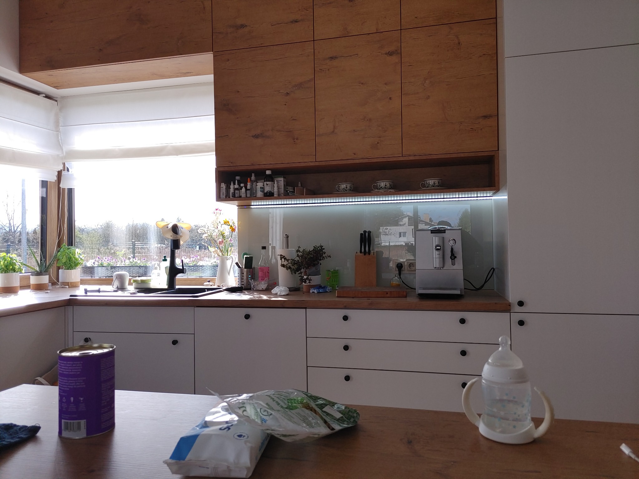Jasna kuchnia z drewnianymi górnymi szafkami, białymi dolnymi szafkami i blatem, ekspres do kawy na blacie, widok na ogród przez okno, na pierwszym planie puszka i butelka dla dziecka.