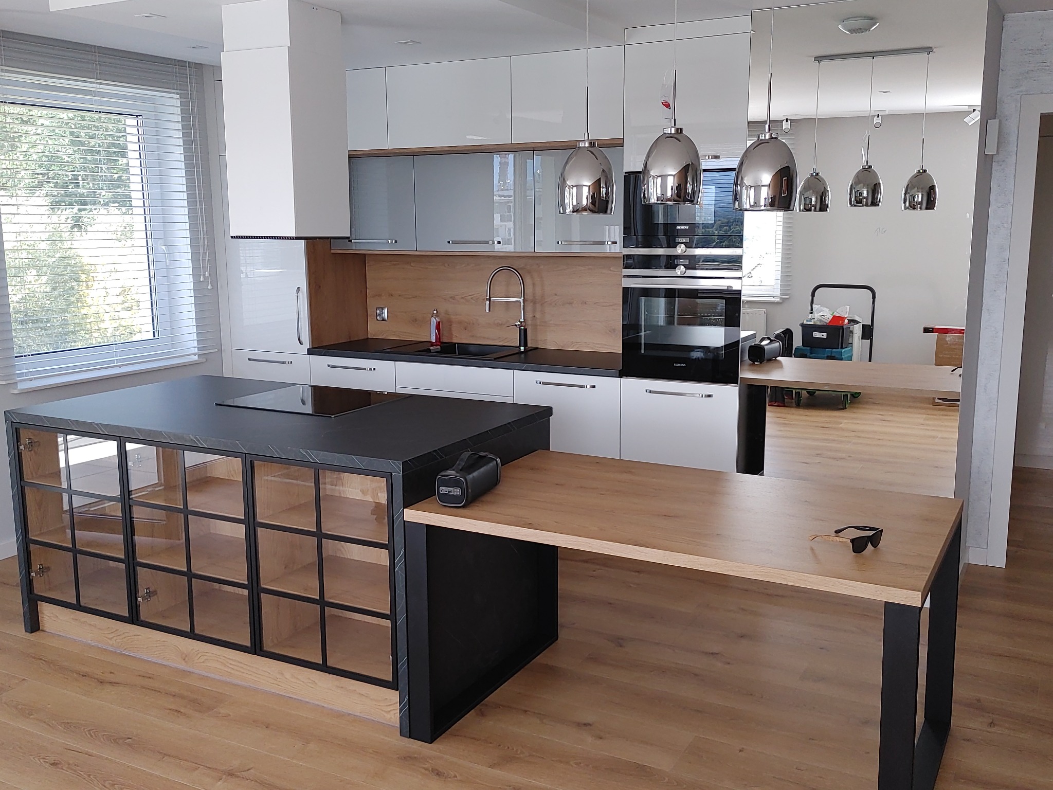 Nowoczesna kuchnia z wyspą i blatem drewnianym, białe szafki wiszące z szarymi frontami, czarny blat, srebrne lampy wiszące, podłoga drewniana, na wyspie leżą okulary przeciwsłoneczne i głośnik.