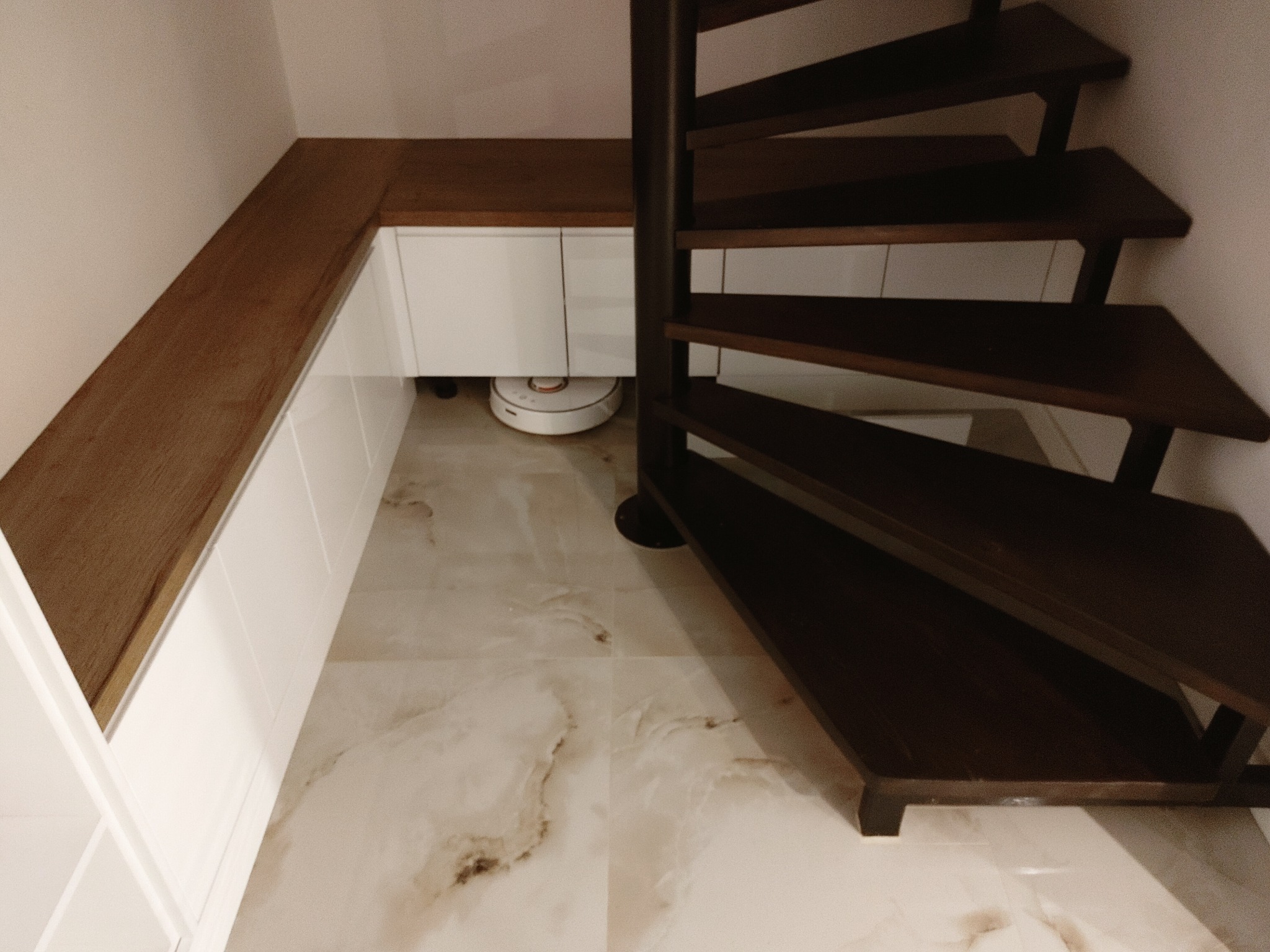 Drewniane schody spiralne w ciemnym odcieniu brązu, obok zabudowy z białymi szafkami i drewnianym blatem, na podłodze marmurkowe płytki. Pod szafkami widoczny robot sprzątający.