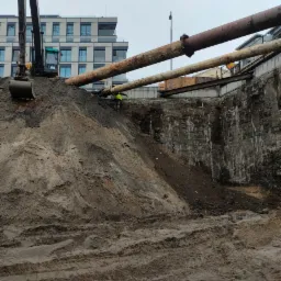 Wykonanie wykopu pod fundamenty budynku z użyciem koparki, widoczne rury i pracownik w kamizelce odblaskowej, teren budowy z ziemią i odsłoniętymi warstwami gruntu.