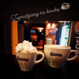 Speedy Romano - Dwie filiżanki kawy z logo 'CoffeeCoffee', jedna z bitą śmietaną, ustawione na barze kawiarni z widocznym wnętrzem i lodówką z napojami w tle.