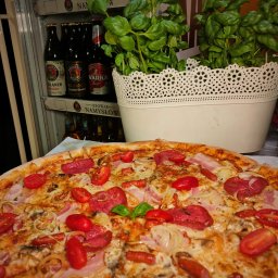 Speedy Romano - Pizza z salami, pomidorkami koktajlowymi, boczkiem i cebulą, z dekoracją w postaci świeżej bazylii, na tle lodówki z piwami i doniczki z bazylią.