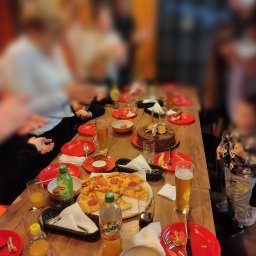 Speedy Romano - Ustawiony stół z pizzą, tortem czekoladowym z gwiazdkami, napojami i czerwonymi talerzami podczas imprezy. Widoczne osoby w tle.