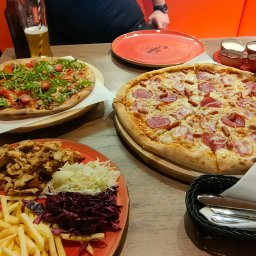 Speedy Romano - Dwa rodzaje pizzy na drewnianych deskach, porcja frytek z mięsem i surówkami na czerwonym talerzu, piwo oraz zestaw sosów na stole w restauracji.