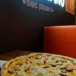 Speedy Romano - Pizza z salami, kukurydzą i papryczkami jalapeno na drewnianym talerzu, w tle neonowy szyld pizzerii z kolorowymi balonami.
