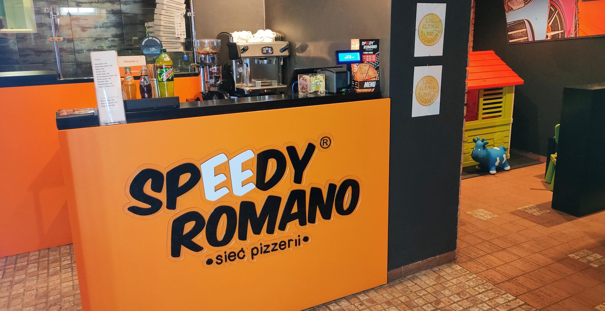 Pomarańczowy bar pizzerii Speedy Romano z ekspresem do kawy, dystrybutorami napojów i menu na ladzie, w tle kącik zabaw dla dzieci z domkiem i niebieskim bujakiem.