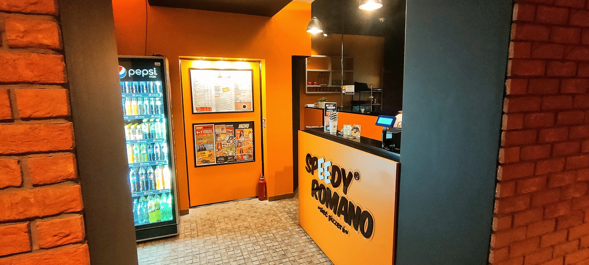 Wnętrze pizzerii Speedy Romano z ladą, menu na ścianie, lodówką z napojami i fragmentami ceglanej ściany.