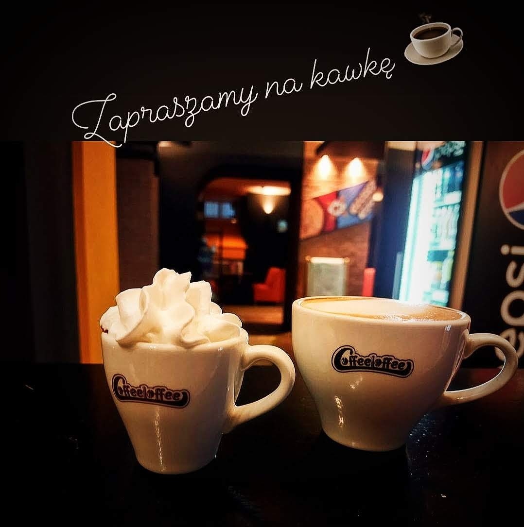 Dwie filiżanki kawy z logo 'CoffeeCoffee', jedna z bitą śmietaną, ustawione na barze kawiarni z widocznym wnętrzem i lodówką z napojami w tle.