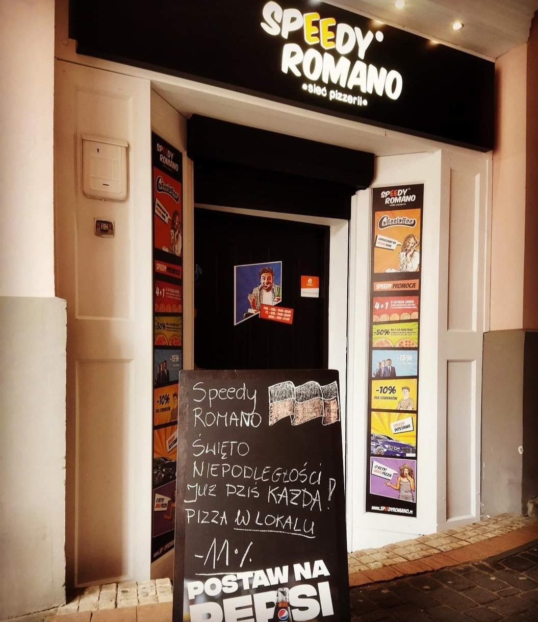 Wejście do pizzerii Speedy Romano z tablicą informującą o promocji z okazji Święta Niepodległości. Na drzwiach i ścianie wiszą plakaty reklamowe z ofertami specjalnymi.
