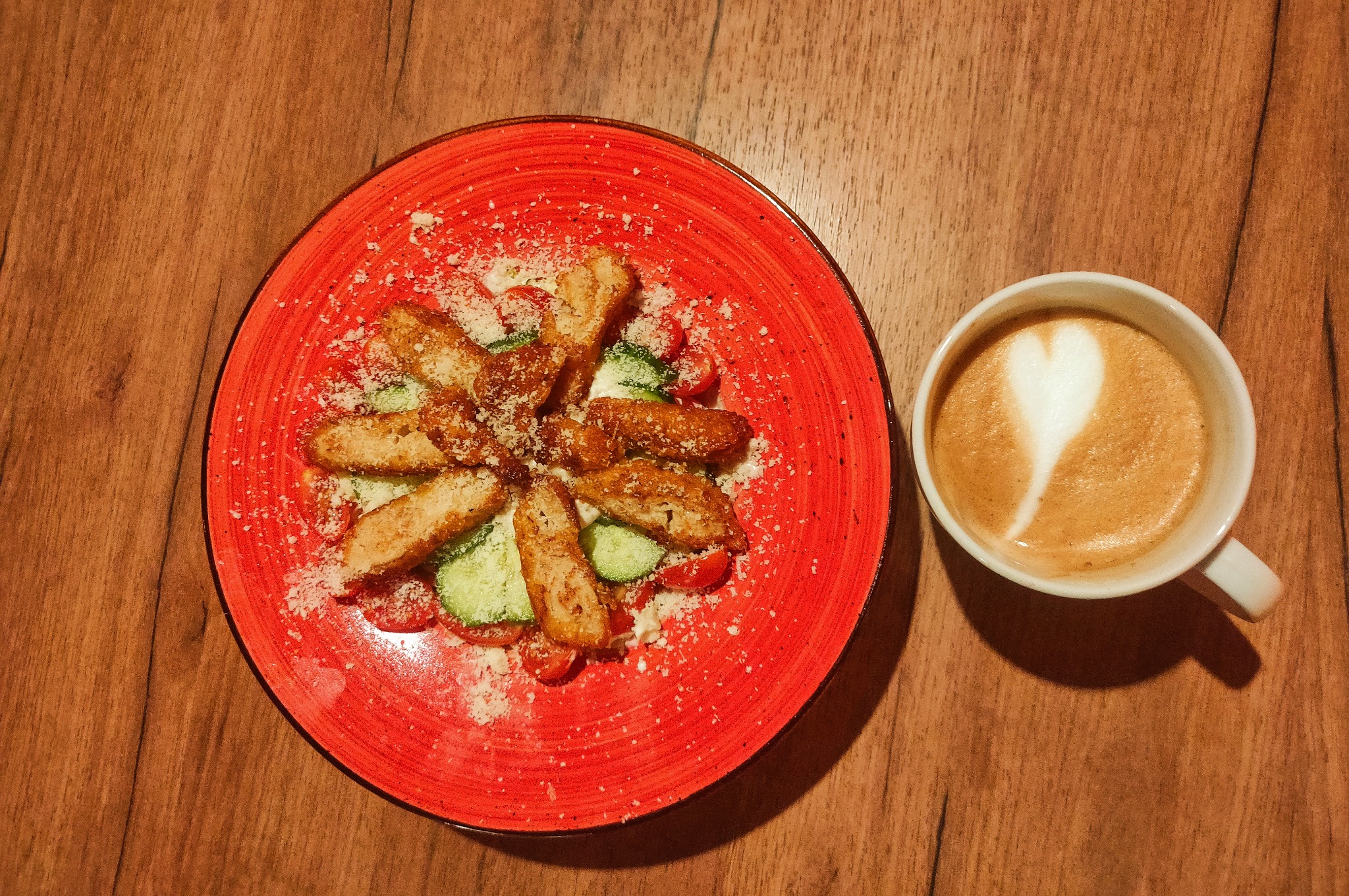 Sałatka z grillowanym kurczakiem, pomidorami, ogórkiem i parmezanem na czerwonym talerzu, obok filiżanka cappuccino z motywem serca.