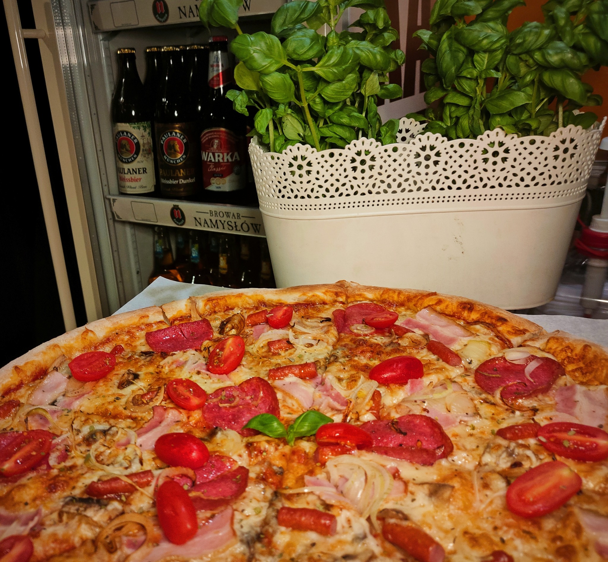 Pizza z salami, pomidorkami koktajlowymi, boczkiem i cebulą, z dekoracją w postaci świeżej bazylii, na tle lodówki z piwami i doniczki z bazylią.