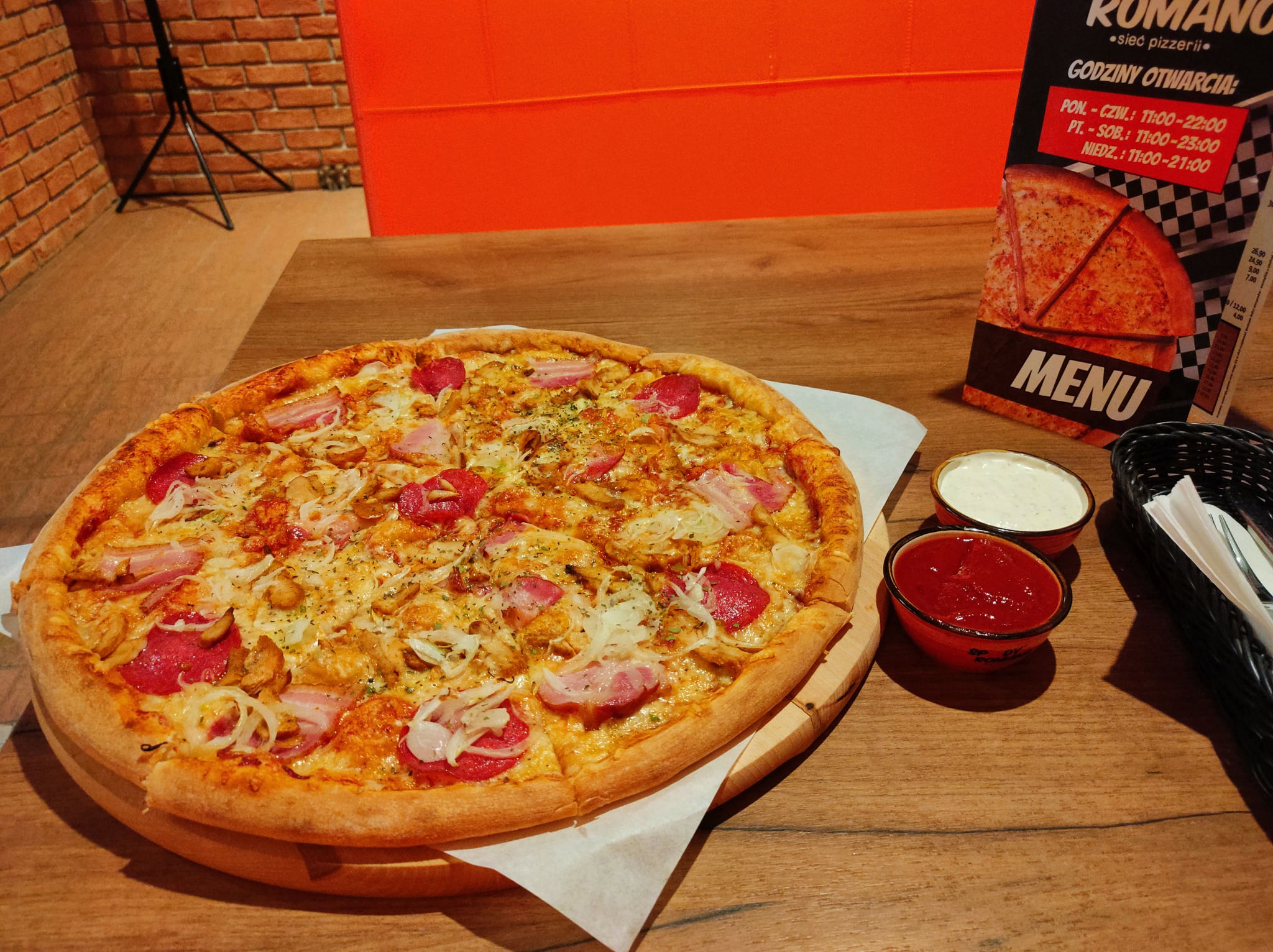 Pizza z salami, boczkiem i cebulą na drewnianym talerzu, serwowana z dwoma sosami w Białymstoku. W tle menu pizzerii Romano.