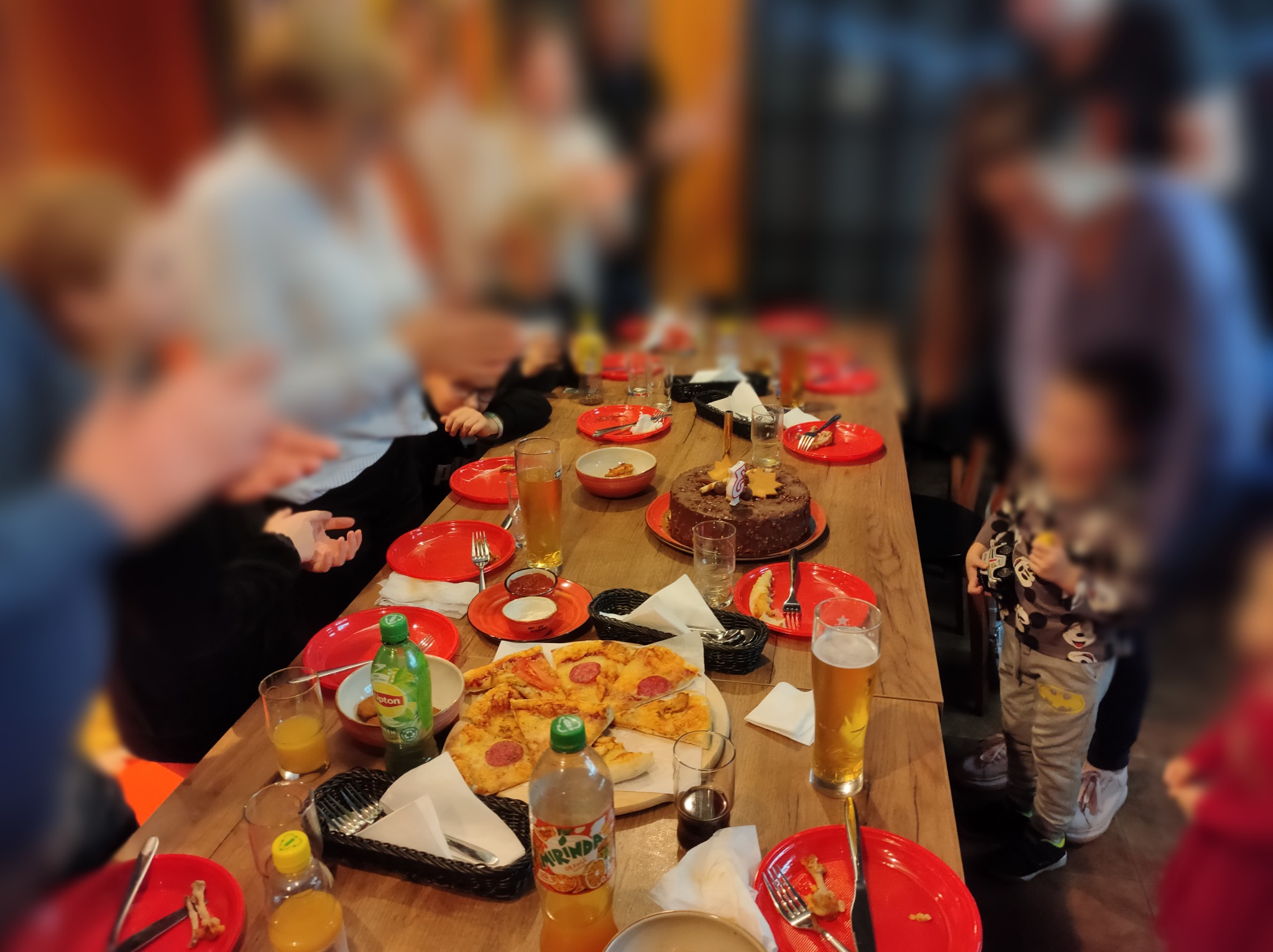 Ustawiony stół z pizzą, tortem czekoladowym z gwiazdkami, napojami i czerwonymi talerzami podczas imprezy. Widoczne osoby w tle.