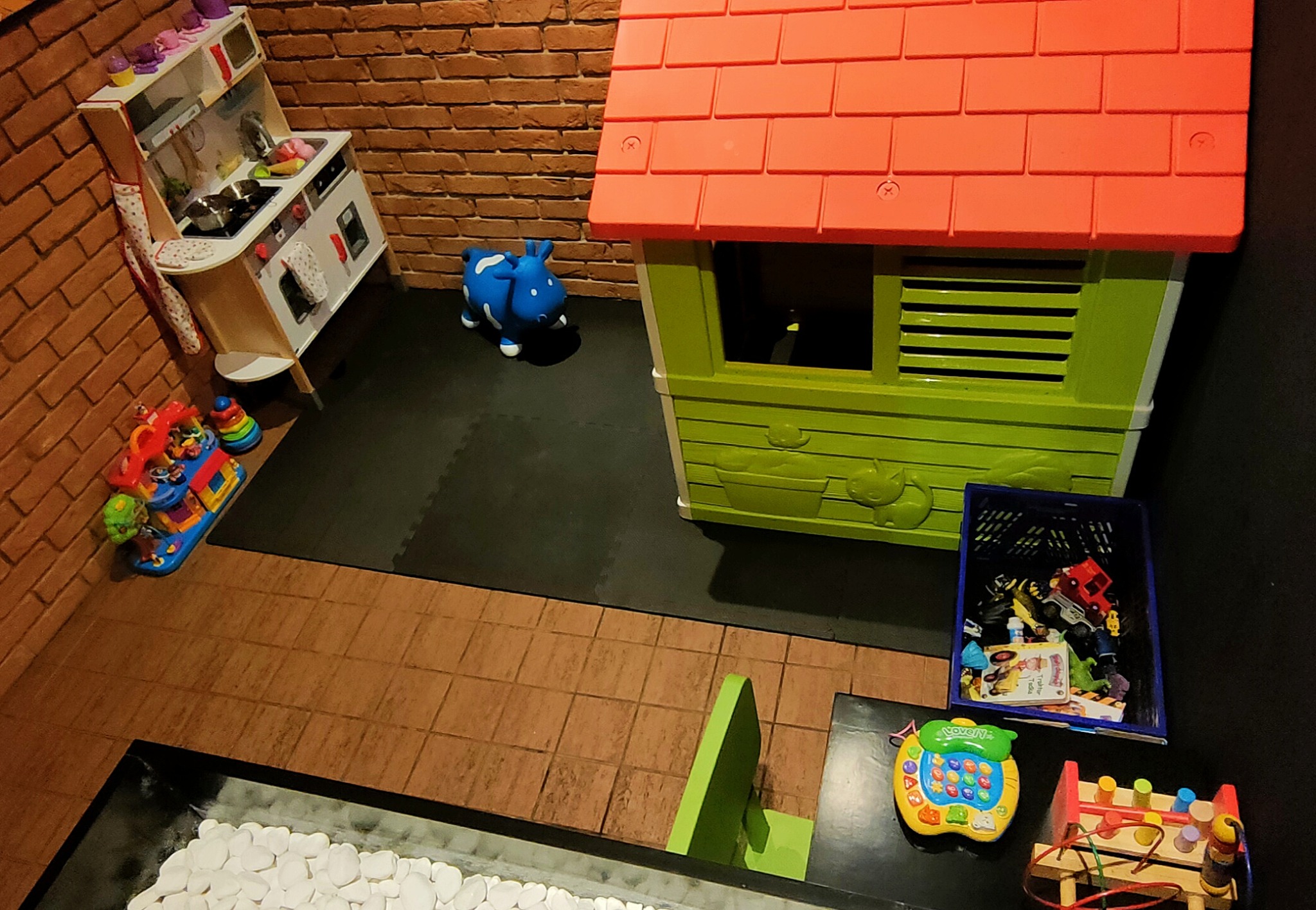 Pokój zabaw dla dzieci z plastikowym domkiem z pomarańczowym dachem, zabawkami i miniaturową kuchnią z ceglanym tłem.