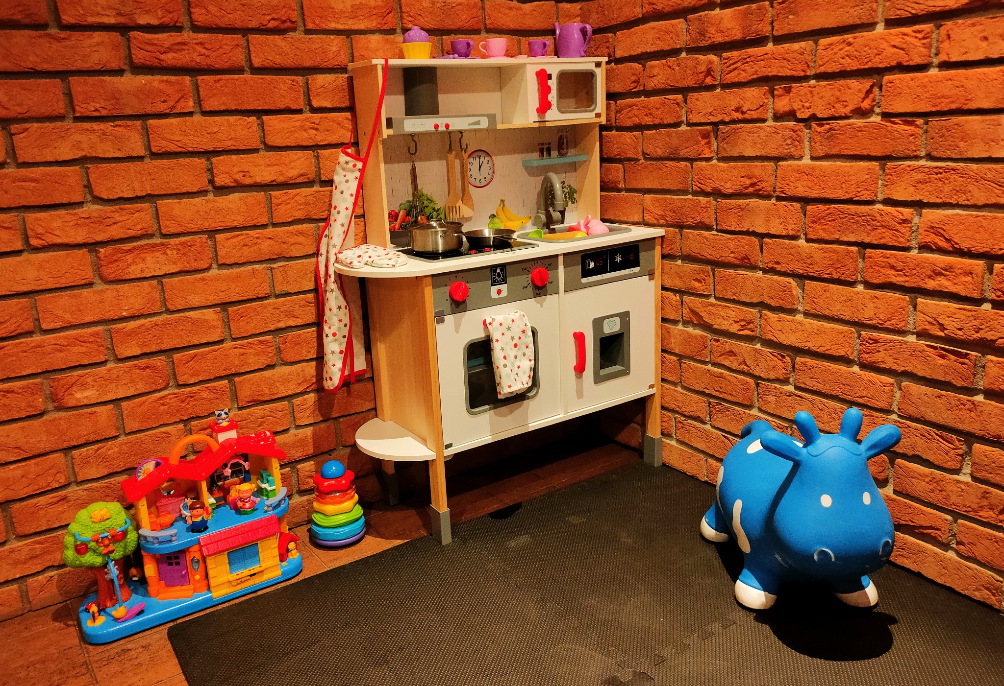 Dziecięca kuchnia zabawkowa z akcesoriami, fartuszkiem i garnkami, ustawiona na tle ściany z czerwonej cegły, obok zabawkowy domek i niebieska dmuchana krówka.