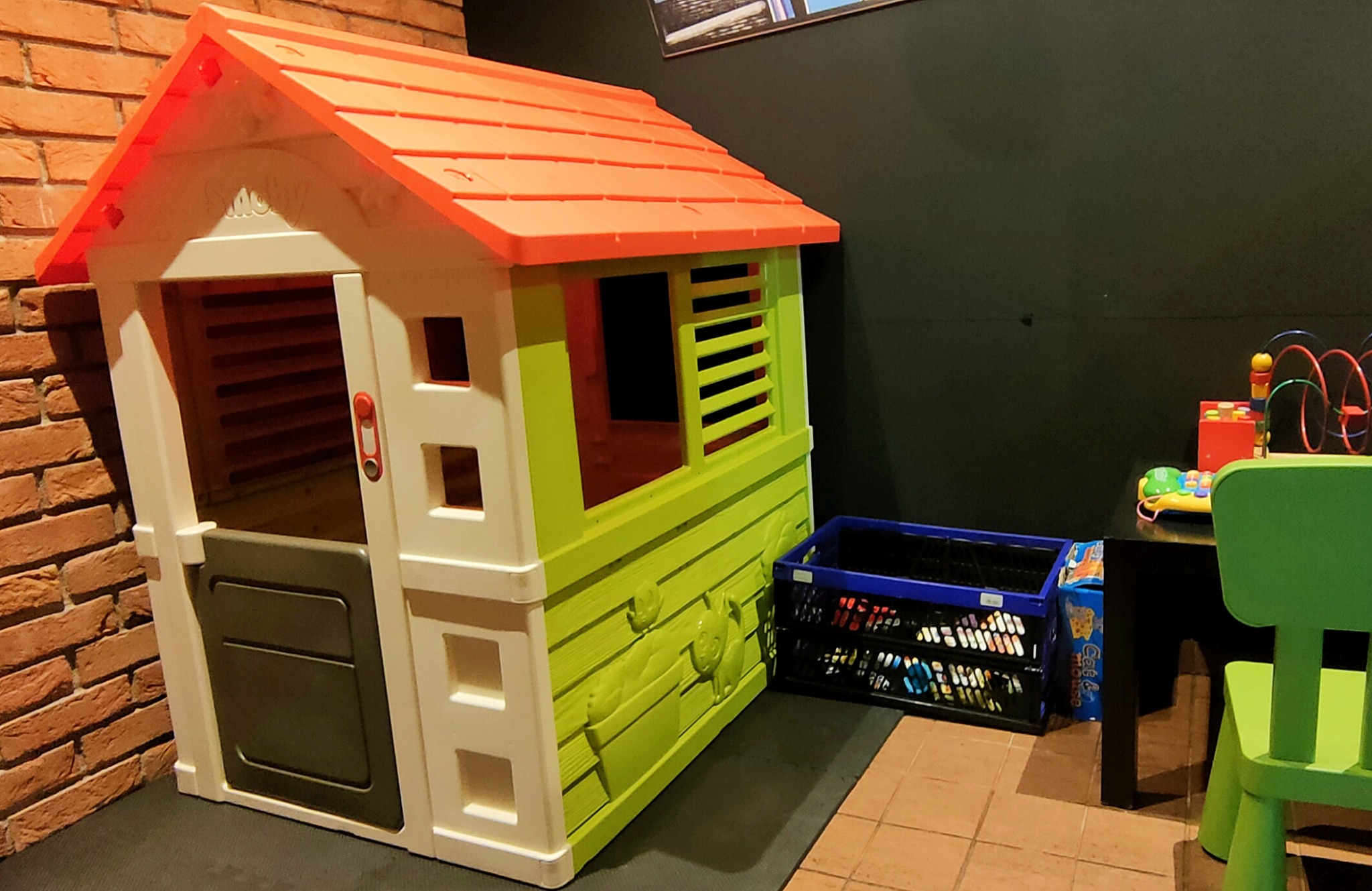 Kolorowy plastikowy domek dla dzieci z pomarańczowym dachem, ustawiony w kąciku zabaw z ceglaną ścianą i czarnym tłem, obok niebieskiego kosza z zabawkami i małego stolika z krzesełkiem.