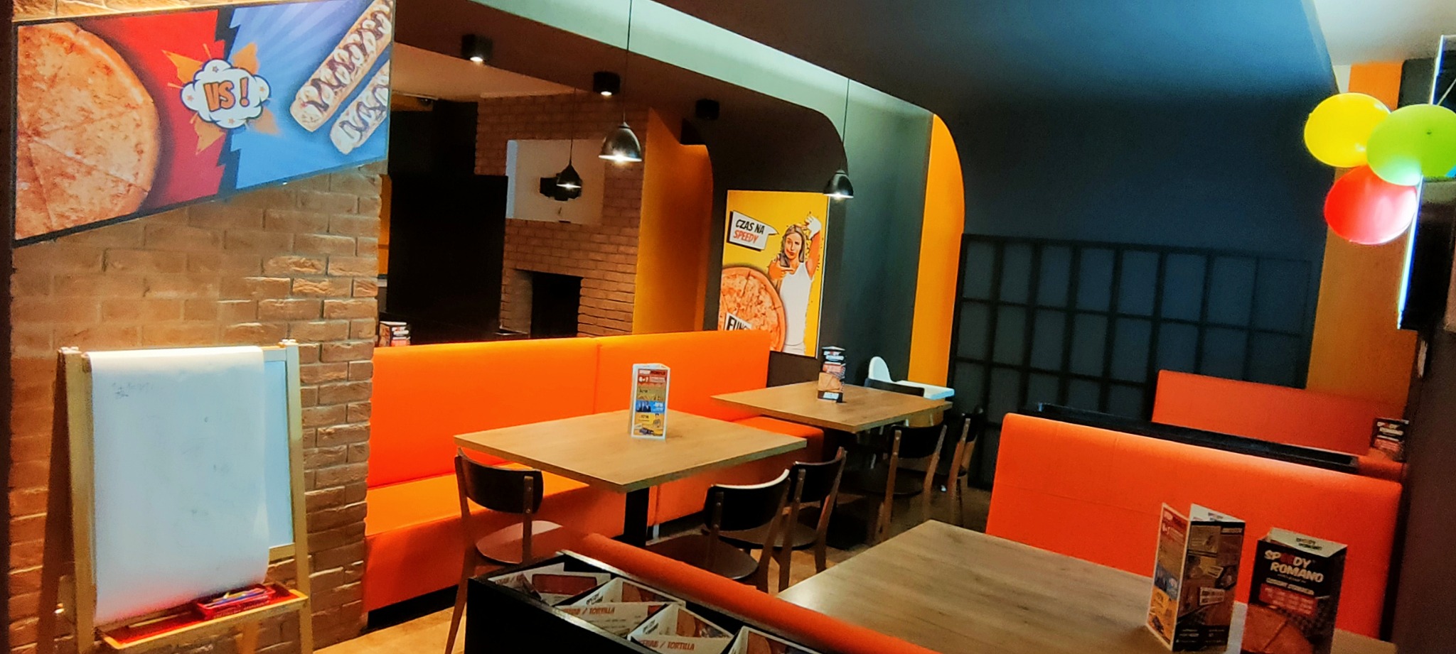 Wnętrze pizzerii z pomarańczowymi kanapami, drewnianymi stolikami i krzesłami, dekoracjami ściennymi z motywem pizzy i balonami.
