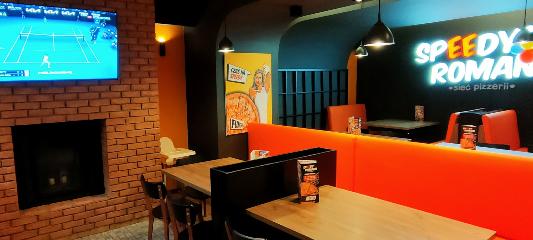 Wnętrze pizzerii z ceglanym kominkiem, telewizorem z meczem tenisowym, neonem 'Speedy Romano' i plakatem reklamowym pizzy.
