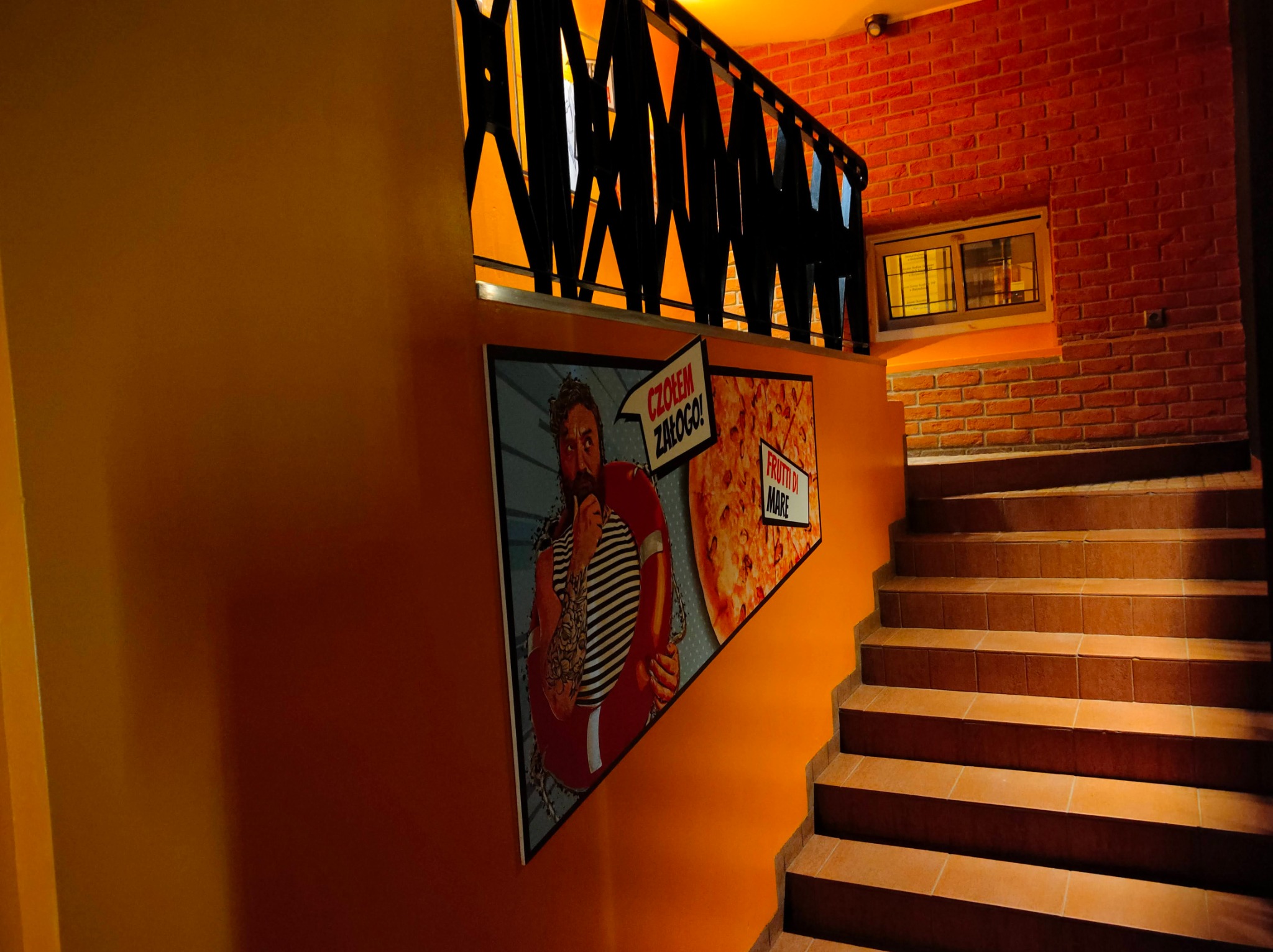 Wnętrze klatki schodowej z pomarańczowymi ścianami, na których wiszą plakaty z motywami marynistycznymi i pizzą Frutti di Mare, schody wyłożone płytkami.