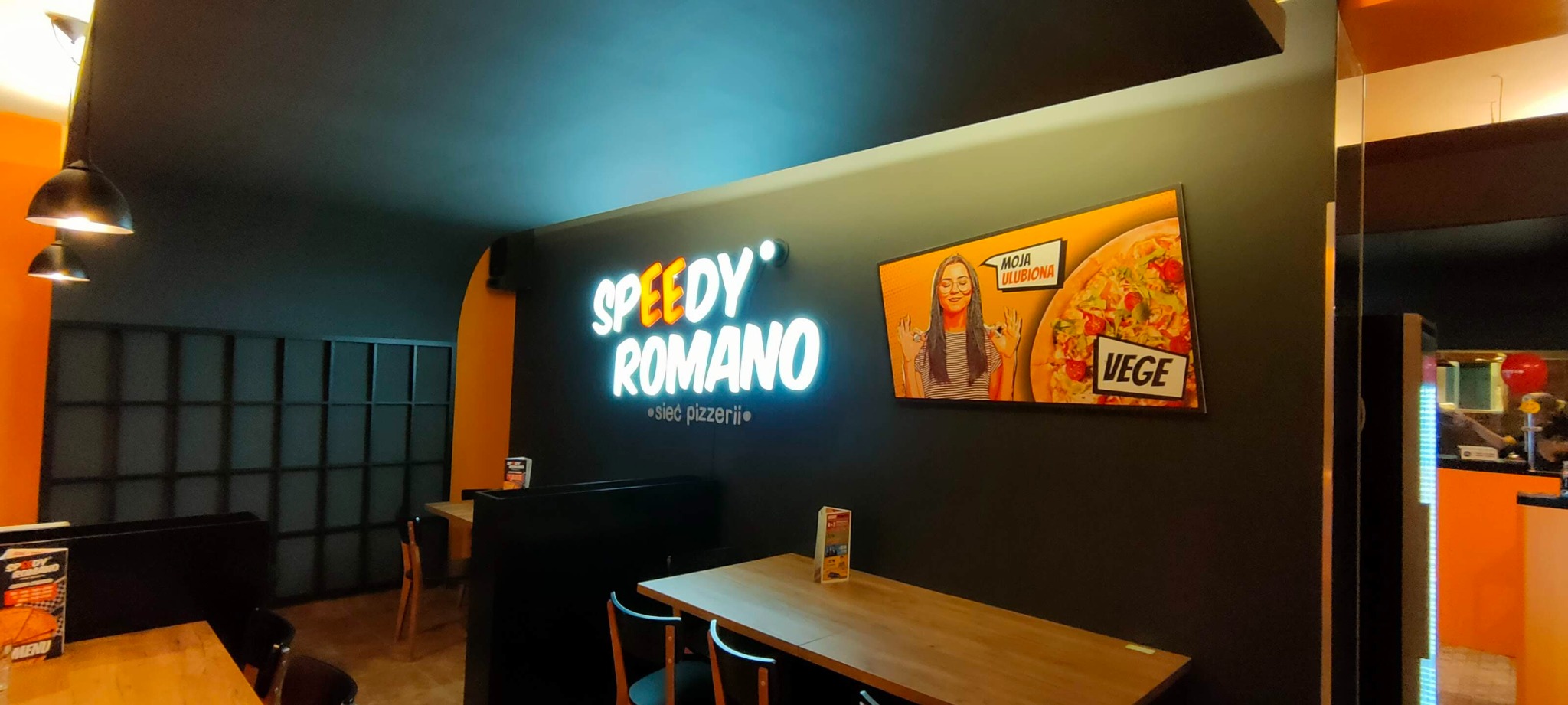 Wnętrze pizzerii, neonowy szyld 'Speedy Romano', plakat z pizzą wege i uśmiechniętą dziewczyną, drewniane stoliki i czarne krzesła, nowoczesny design.