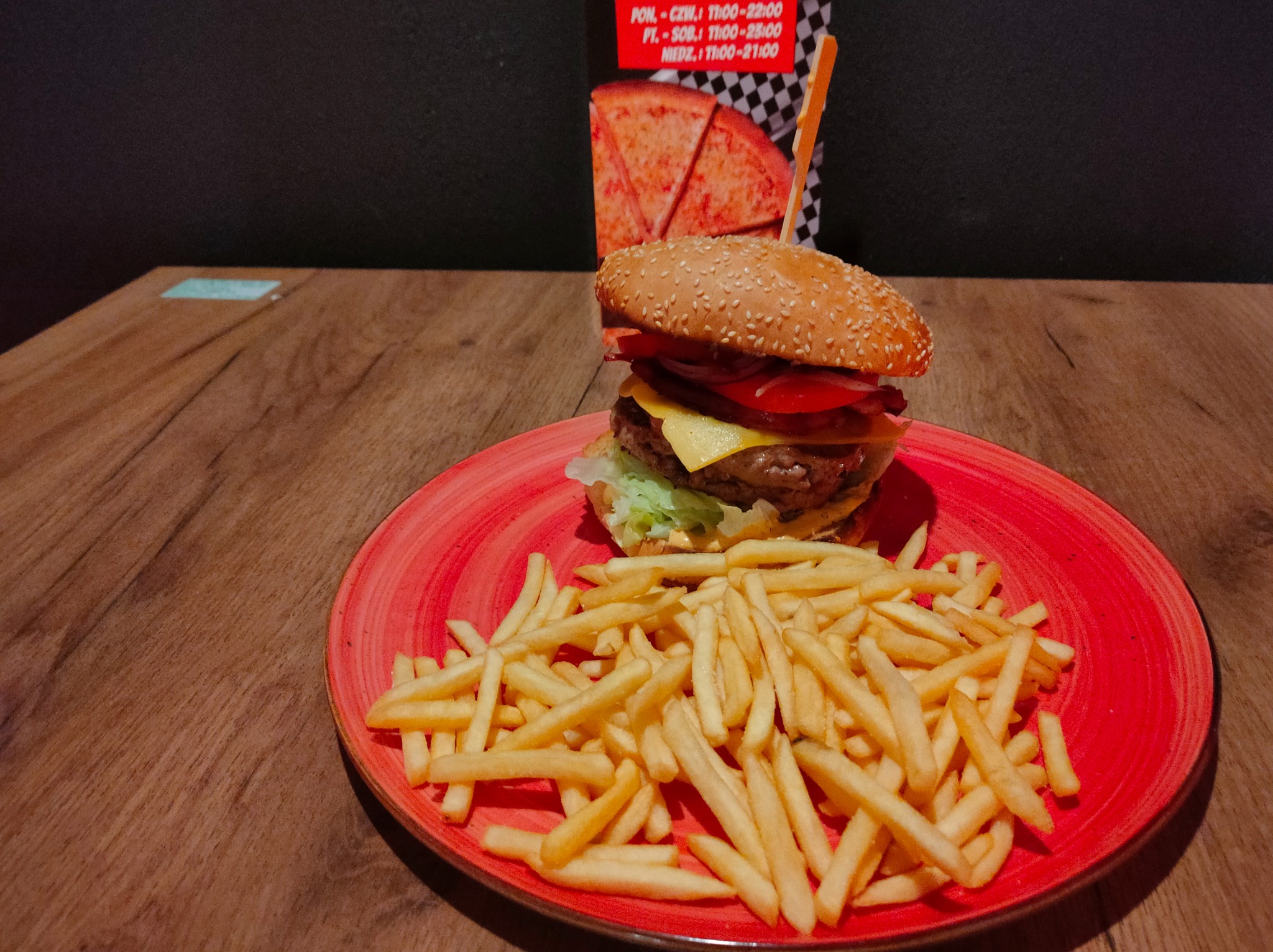 Burger z wołowiną, serem, warzywami i frytkami na czerwonym talerzu, z menu restauracji w tle.