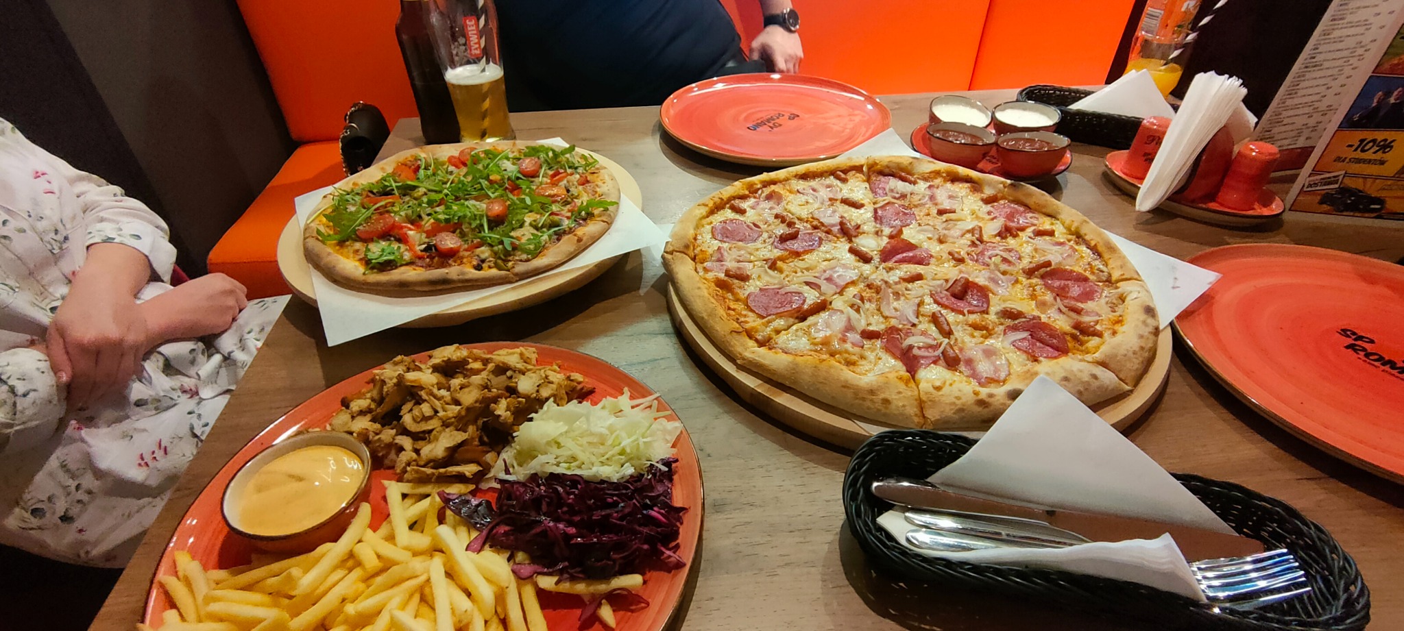 Dwa rodzaje pizzy na drewnianych deskach, porcja frytek z mięsem i surówkami na czerwonym talerzu, piwo oraz zestaw sosów na stole w restauracji.