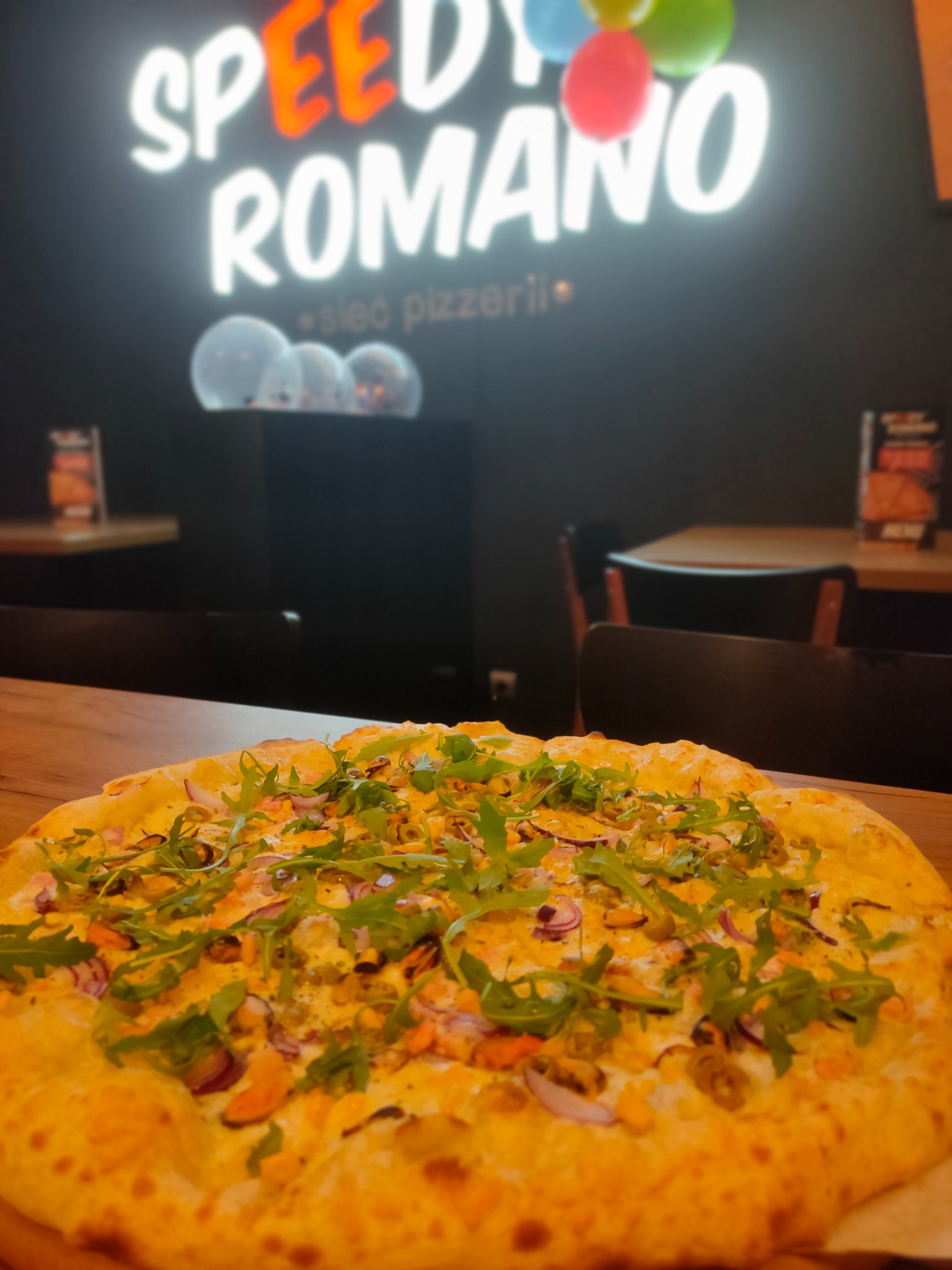 Pizza z owocami morza i rukolą na drewnianym stole, w tle neonowy szyld pizzerii Speedy Romano i dekoracyjne balony.