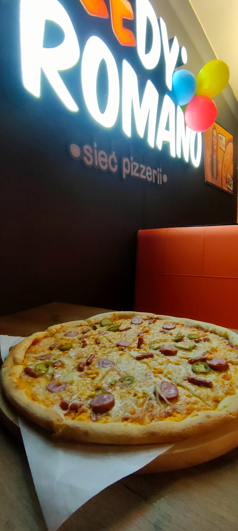 Pizza z salami, kukurydzą i papryczkami jalapeno na drewnianym talerzu, w tle neonowy szyld pizzerii z kolorowymi balonami.
