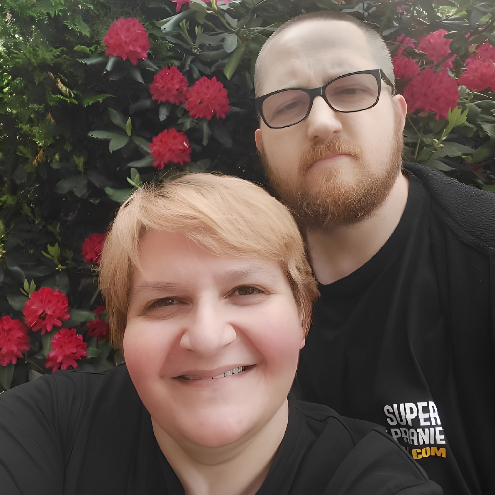 Uśmiechnięta kobieta i mężczyzna w okularach, pozujący do zdjęcia na tle kwitnącego krzewu różaneczników; kobieta ma na sobie czarną koszulkę z napisem 'SUPER PRANIE .COM'.