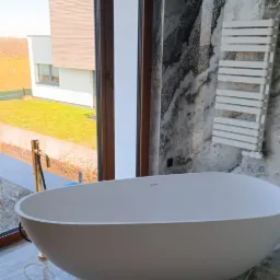 Nowoczesna łazienka z wolnostojącą wanną o owalnym kształcie, złota bateria podłogowa, marmurowe ściany i podłoga, grzejnik drabinkowy oraz widok na ogród przez duże okno.