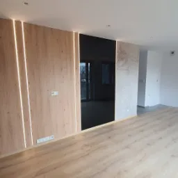 Nowoczesne wnętrze: ściana z paneli drewnopodobnych z podświetleniem LED, czarna tafla lustra, podłoga z paneli, ściana z delikatnym wzorem, wisząca lampa, widoczne rury CO.