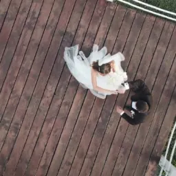 Widok z góry na parę młodą na drewnianym pomoście, otoczonym zielenią i białymi linami, uchwycony dronem.