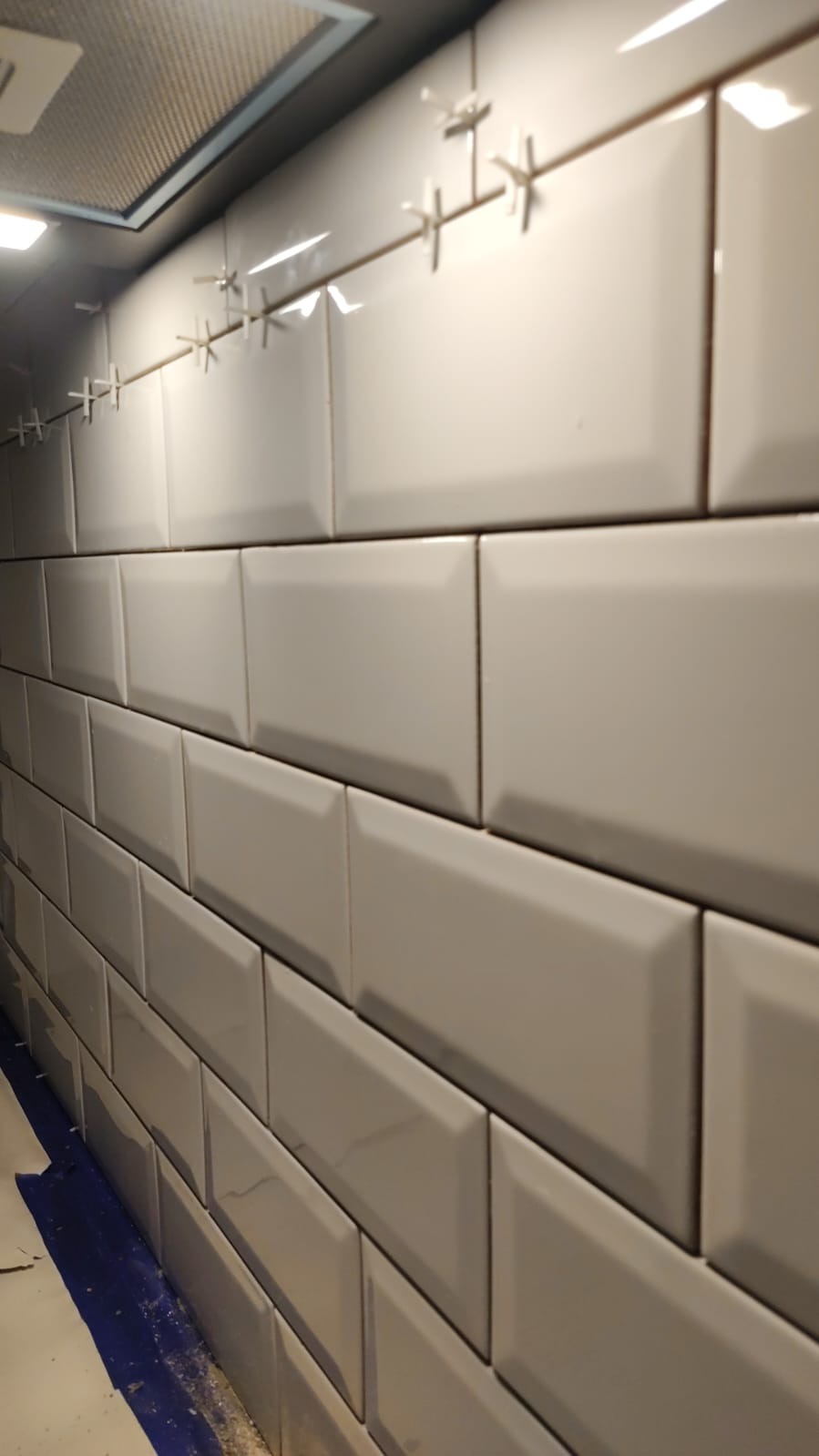 Świeżo położone, błyszczące, beżowe płytki typu metro na ścianie, z widocznymi krzyżykami dystansowymi i zabezpieczoną folią podłogą.