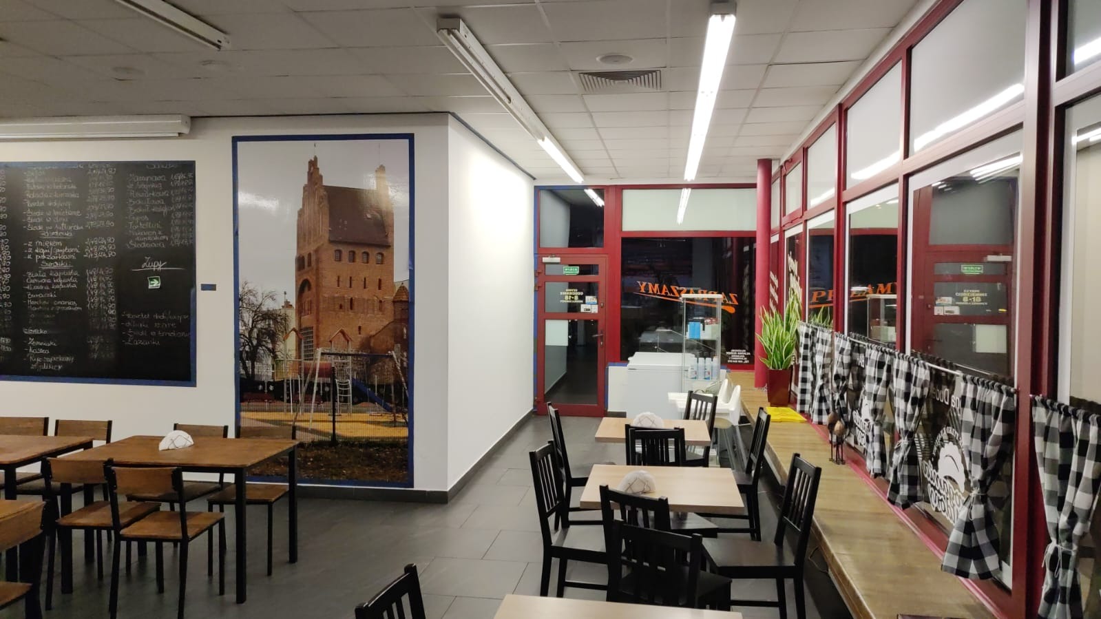 Wnętrze lokalu gastronomicznego z drewnianymi stolikami i krzesłami, tablicą z menu na ścianie oraz dużym zdjęciem zamku i placu zabaw.