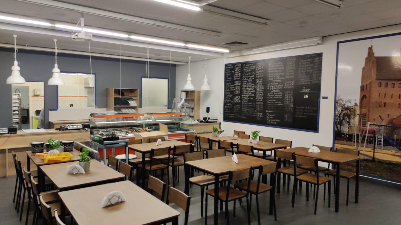 Wnętrze lokalu gastronomicznego z drewnianymi stołami i krzesłami, tablicą z menu na ścianie i witryną chłodniczą w tle.