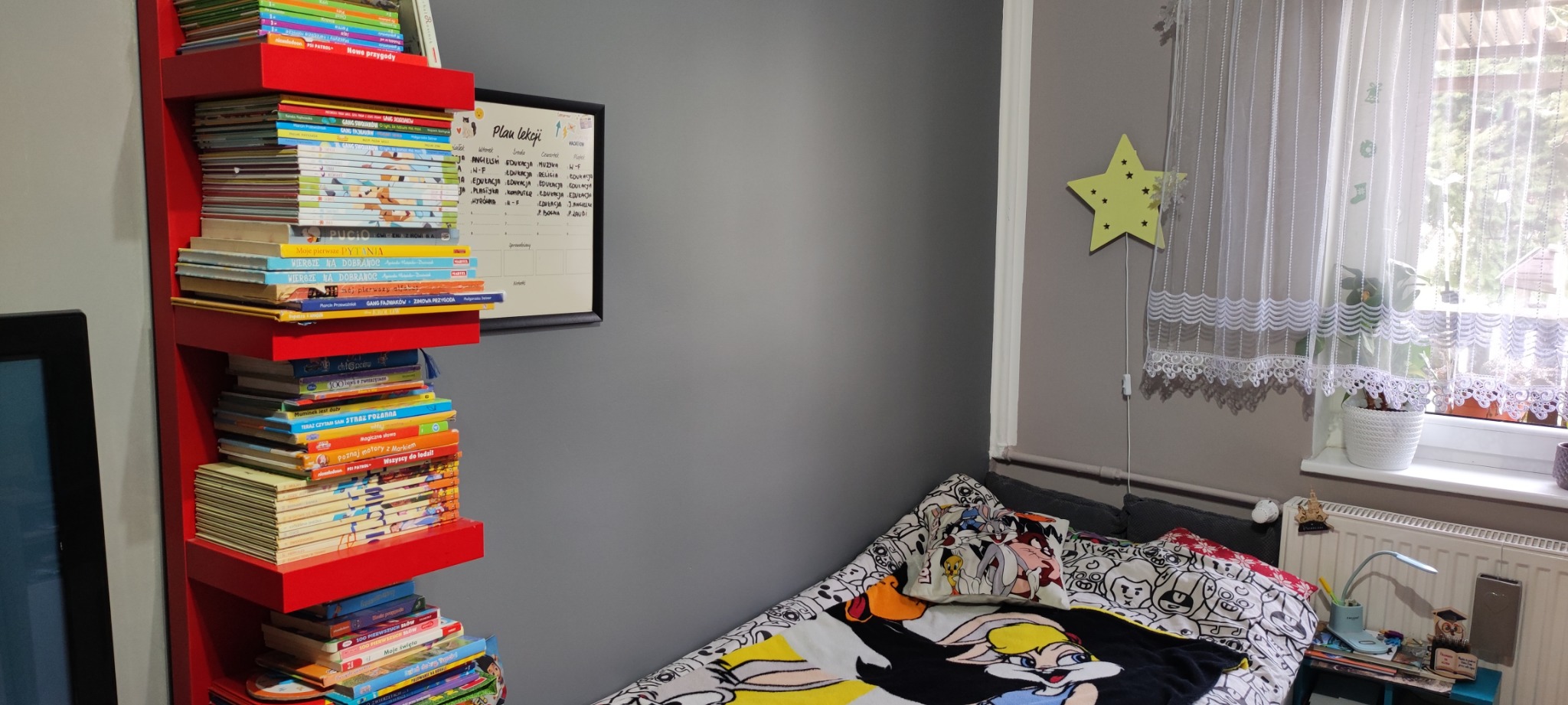 Pokój dziecięcy z czerwonym regałem pełnym książek, planem lekcji na ścianie i łóżkiem z pościelą w kreskówki.