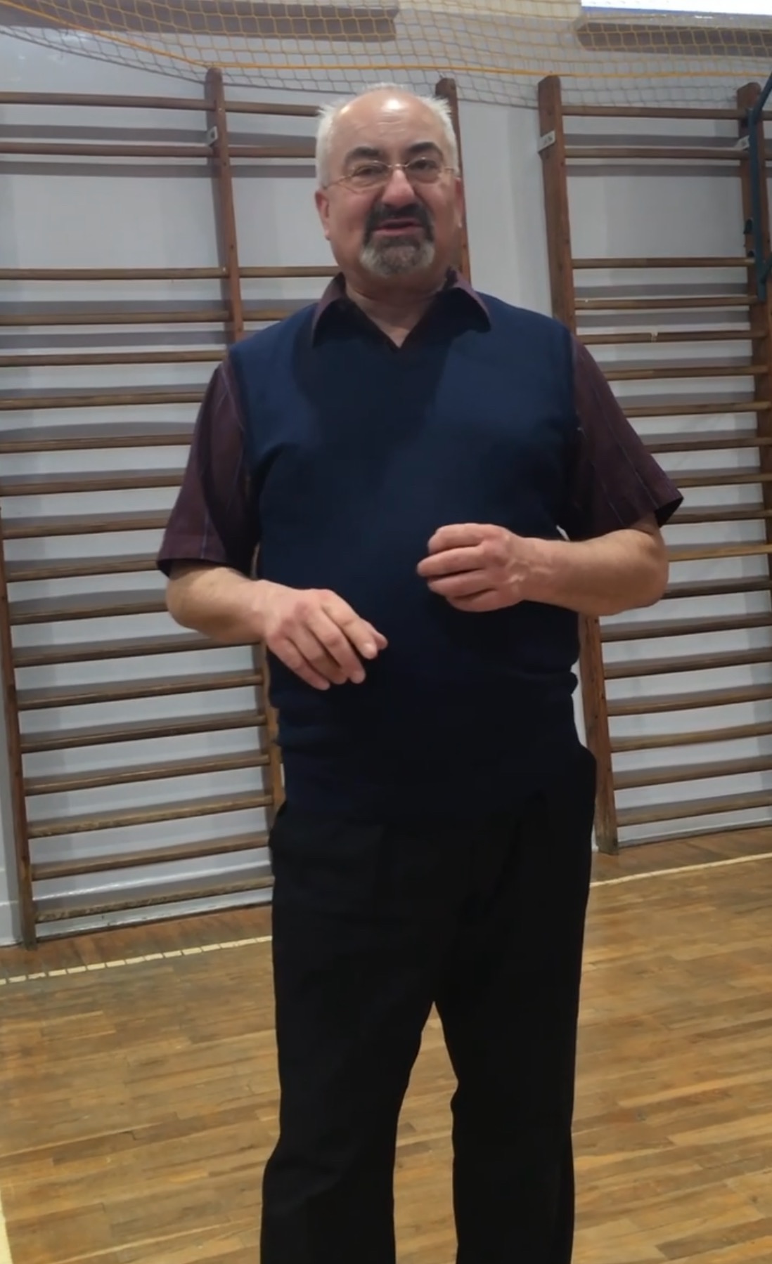 Instruktor tańca w okularach, prezentujący gestykulację dłoni w sali z drabinkami gimnastycznymi.