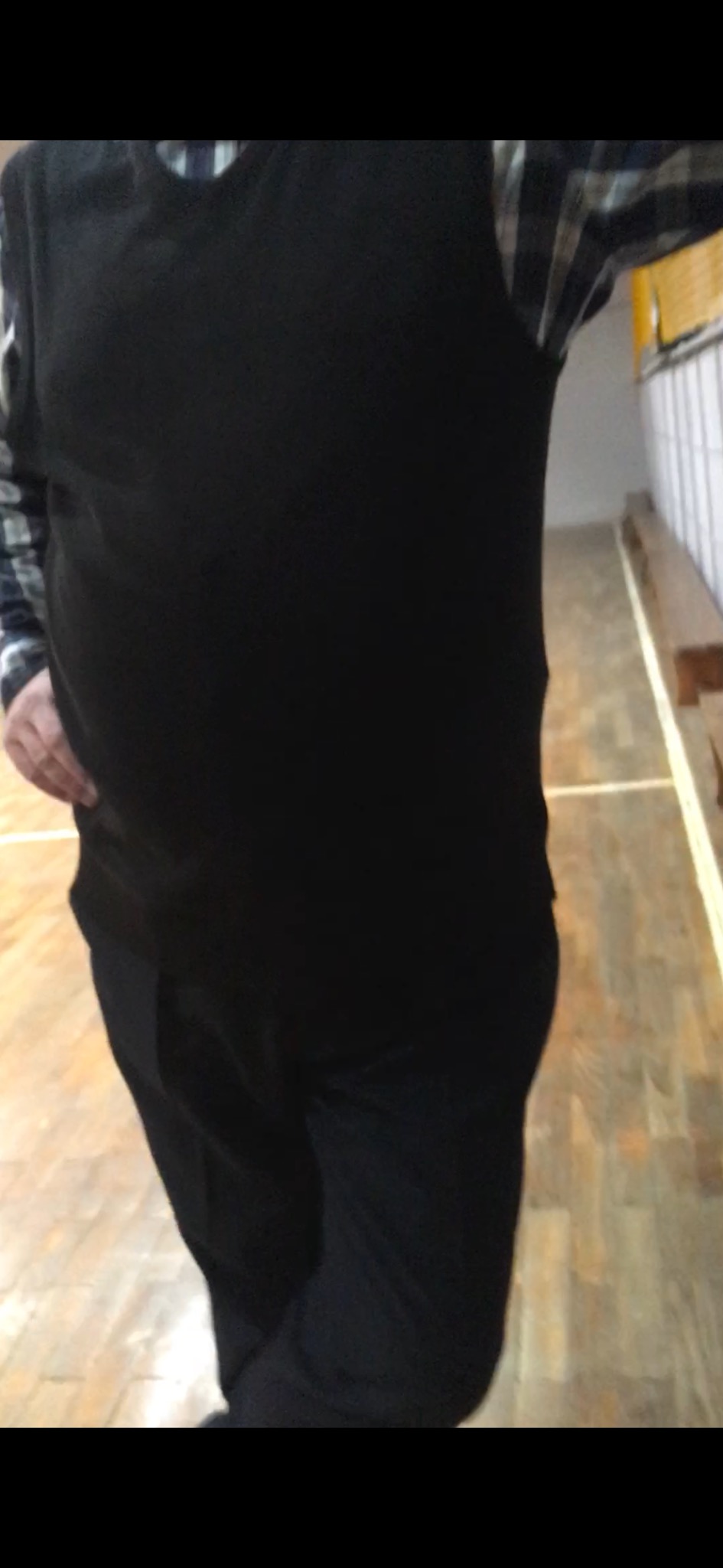 Mężczyzna w czarnej kamizelce i spodniach, widziany od pasa w dół, stojący na drewnianej podłodze w pomieszczeniu z białą ścianą i drewnianą ławką.