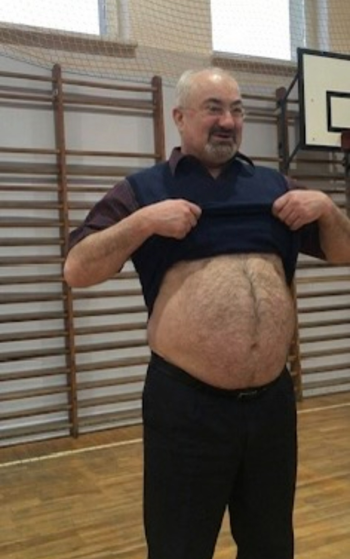 Mężczyzna z brodą, w czarnych spodniach i kamizelce, podnosi koszulkę, ukazując brzuch w sali gimnastycznej z drabinkami i koszem do koszykówki w tle.