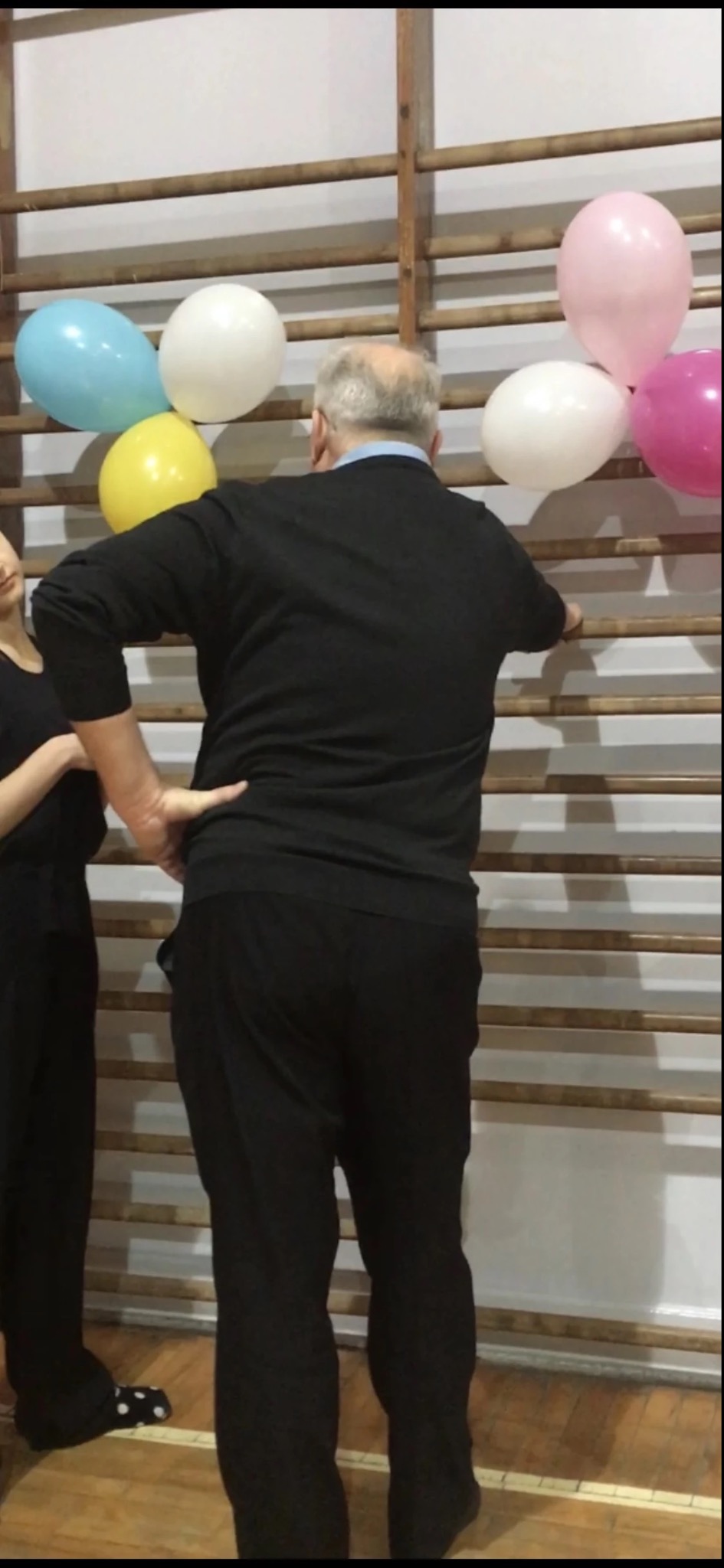 Mężczyzna w czarnym stroju opiera się o drabinki gimnastyczne udekorowane kolorowymi balonami, obok stoi młodsza osoba w czarnym stroju z ręką na biodrze, widoczne stopy w skarpetach w kropki.