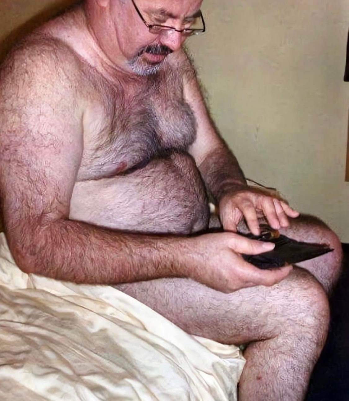 Mężczyzna z okularami siedzi na łóżku i ogląda coś na czarnym urządzeniu elektronicznym w dłoniach.