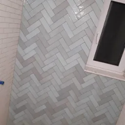 Świeżo ułożona, połyskująca glazura w odcieniach szarości i bieli, ułożona we wzór jodełki na ścianie łazienki, z widocznym oknem w górnym rogu i fragmentem drewnianej podłogi w dolnym rogu.