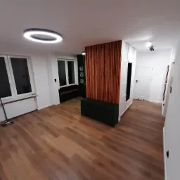 Jasne, przestronne wnętrze po remoncie z drewnianą podłogą, białymi ścianami i zabudową meblową z drewnianymi lamelami oraz nowoczesnym oświetleniem sufitowym.