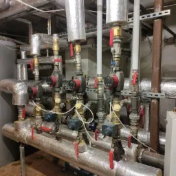 Rozbudowana instalacja grzewcza z pompami obiegowymi, zaworami i izolowanymi rurami w kotłowni, widoczne elementy kotła gazowego z żółtym akcentem.