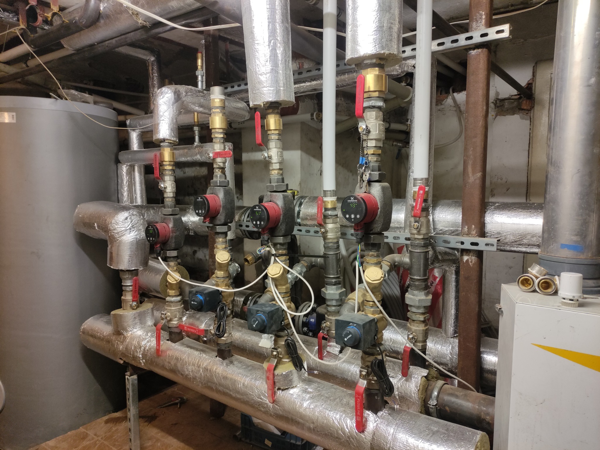 Rozbudowana instalacja grzewcza z pompami obiegowymi, zaworami i izolowanymi rurami w kotłowni, widoczne elementy kotła gazowego z żółtym akcentem.