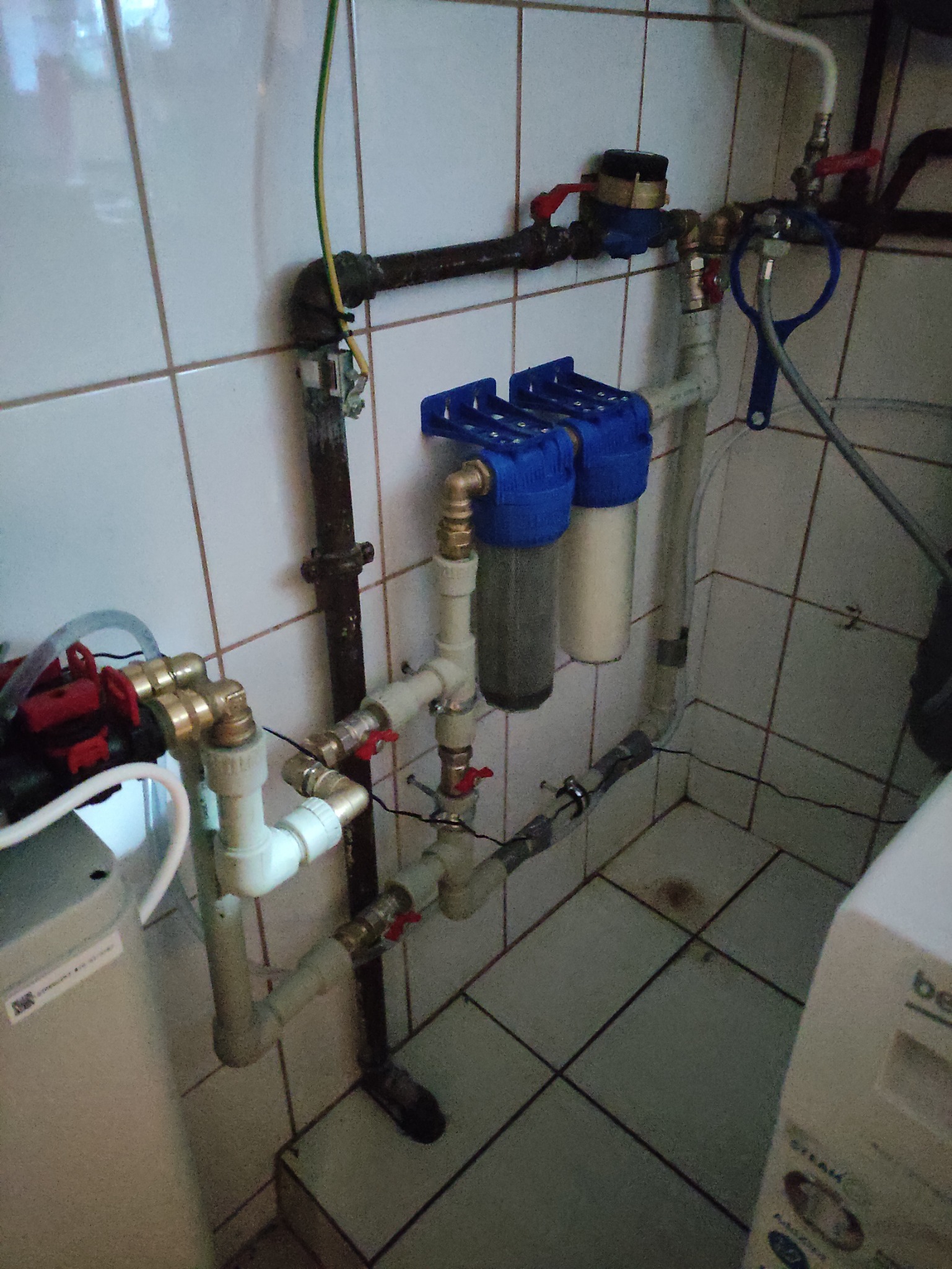 Kompleksowa instalacja wodno-kanalizacyjna z filtrami mechanicznymi, licznikiem i zaworami odcinającymi, widoczna na tle białych płytek.