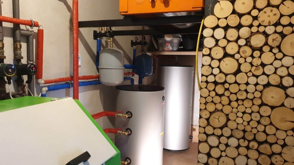 Kotłownia z dwoma srebrnymi zbiornikami buforowymi, rurami w izolacji termicznej i dekoracyjną ścianą z przekrojów drewna.