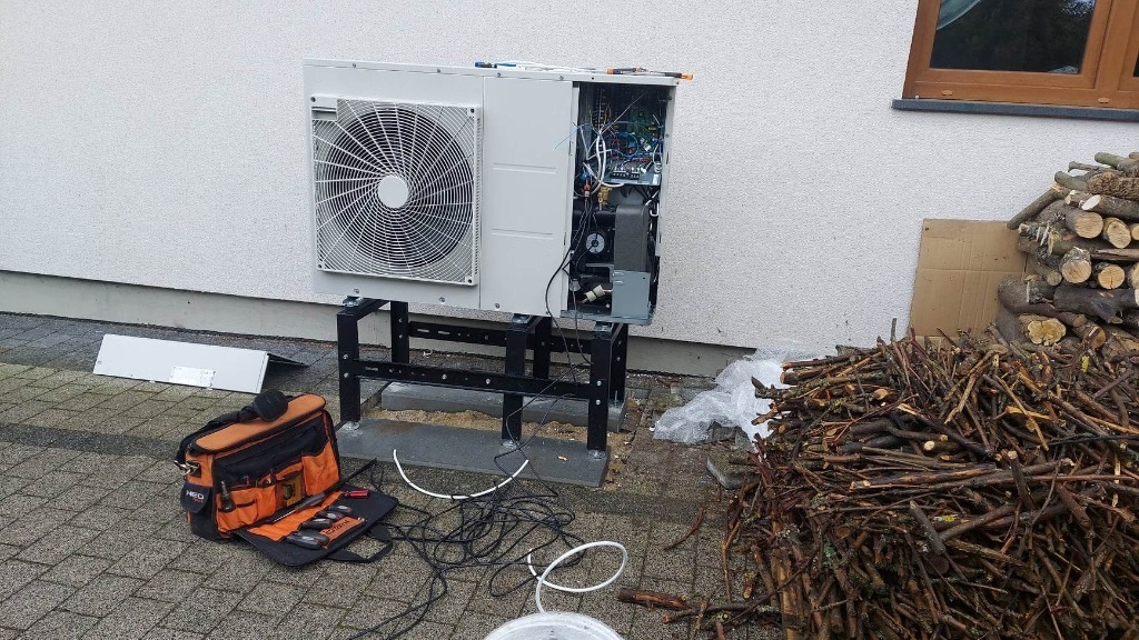 Zewnętrzna jednostka pompy ciepła na czarnym stelażu, z otwartą obudową ukazującą wewnętrzne komponenty, obok leży torba z narzędziami, a w tle widać stos drewna.